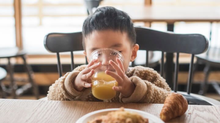 小男孩坐在桌旁吃早餐和喝饮料，喝着一杯橙汁