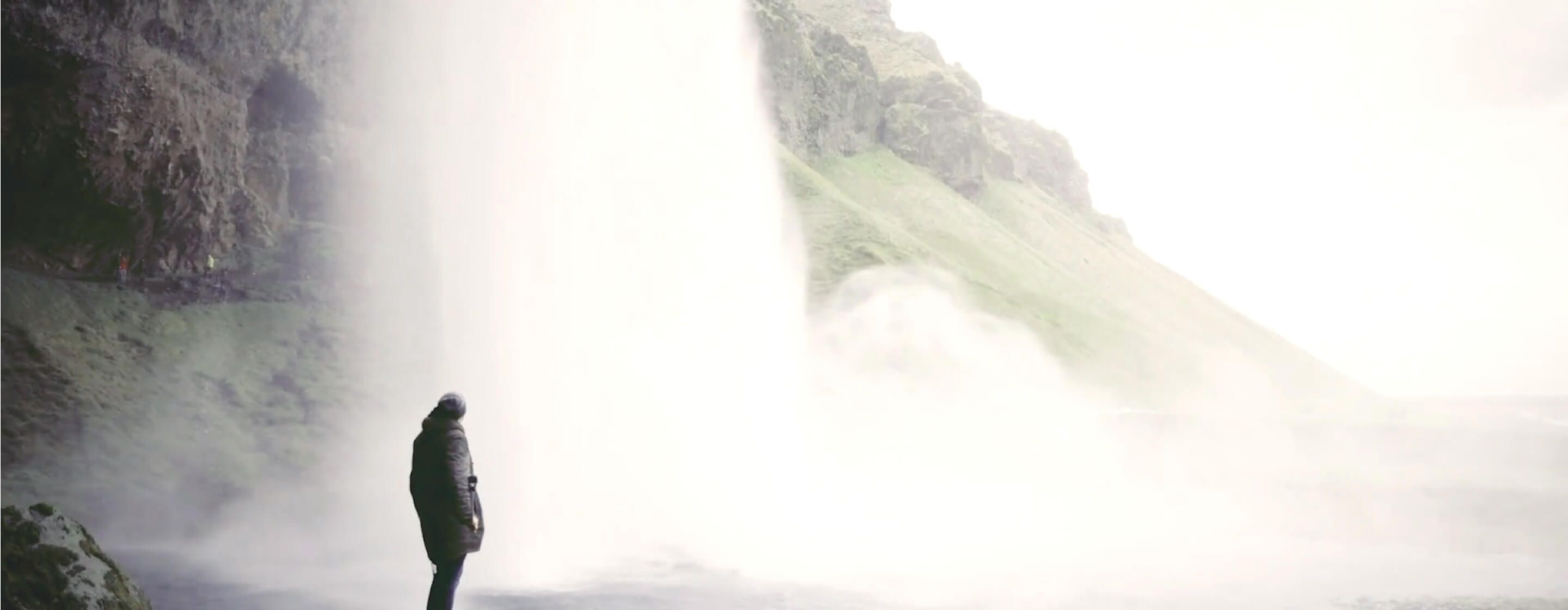 Person in a coat standing next to a massive cascading waterfall with rocks and green hills behind