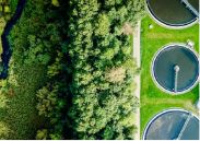 Aerial view of a municipal water treatment plant's settling tanks next to a dense bank of trees and a brook