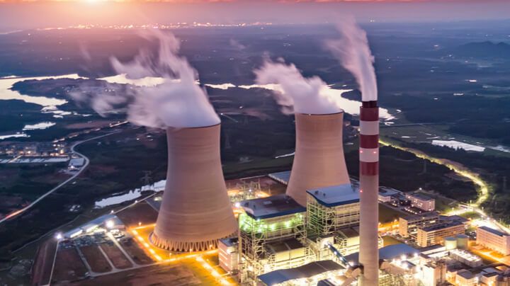 Aerial view of a brightly lit nuclear power generation plant at sunset, with a river and city in the distance