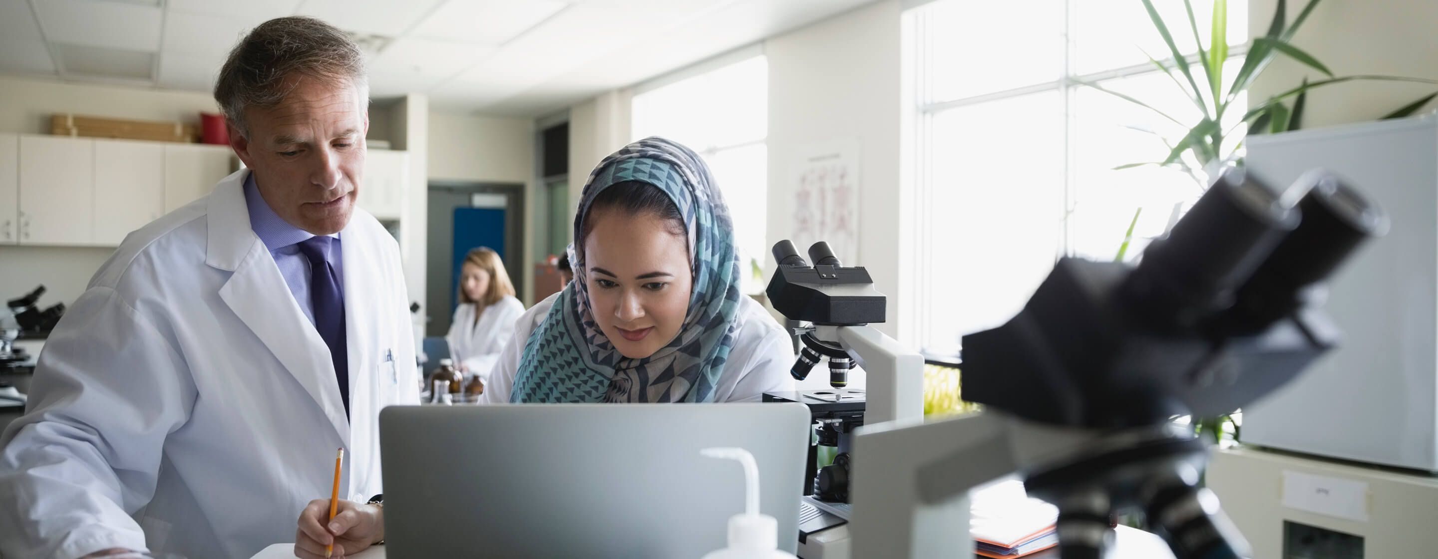 Woman and man wearing white lab coats working at a computer in a laboratory with microscopes and another woman behind them 