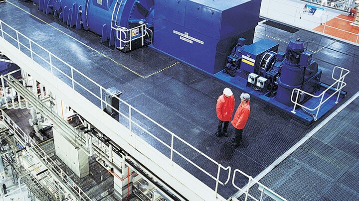 Overhead view of two people in hard hats and orange jackets talking on the top floor of a large multistory industrial facility.