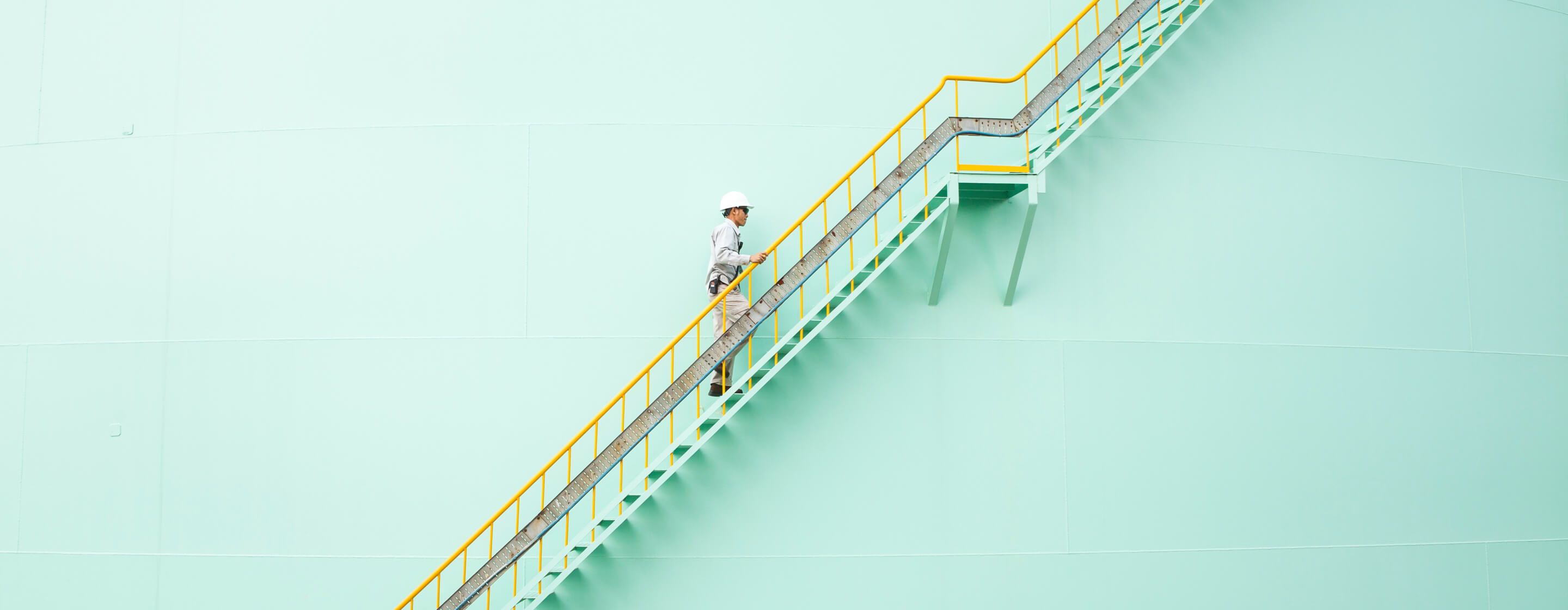 Worker wearing a hardhat climbing a steep staircase on the side of a massive lightgreen chemical and petrochemical tank