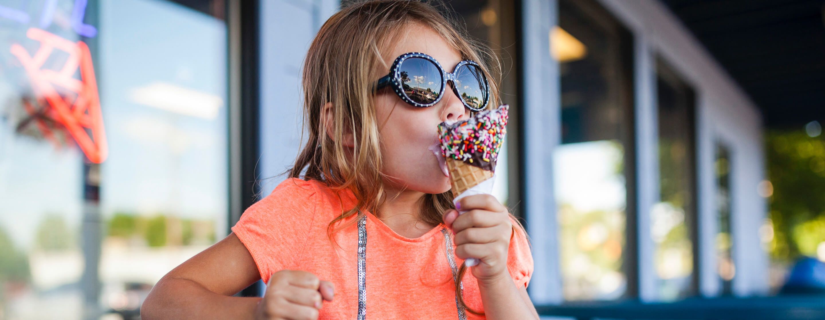 Young girl in sunglasses eating a chocolate and sprinkles-covered ice cream cone in front of an ice cream shop
